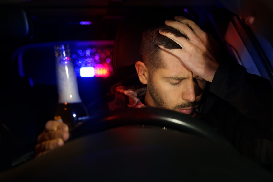 Driver holding his head in distress while sitting behind the wheel with a beer bottle, surrounded by police lights, representing the aftermath of a drunk driving accident with injuries.